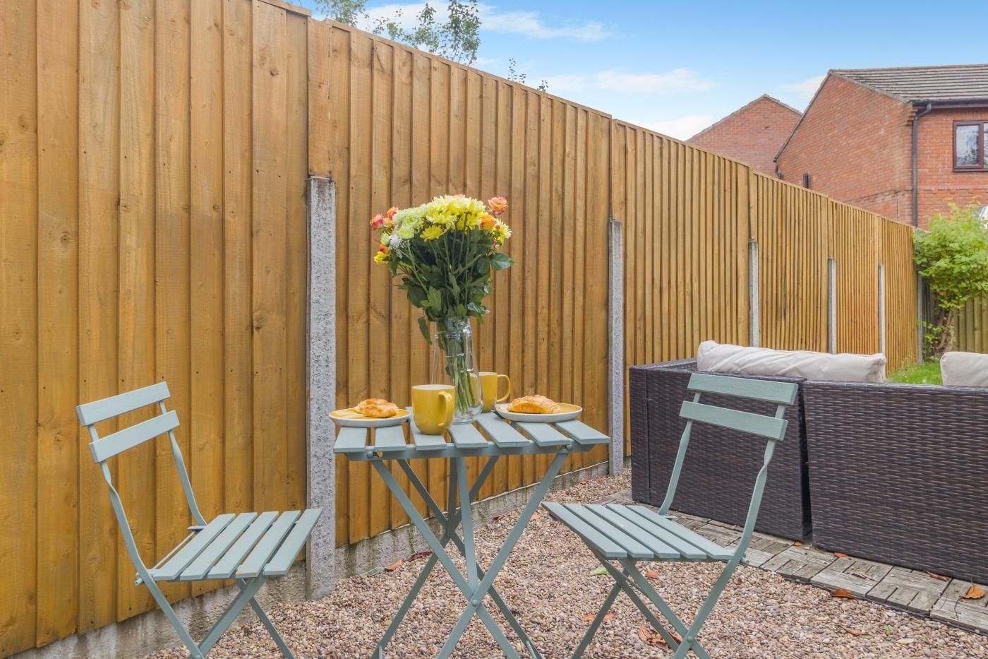 A patio area with a wooden fence, a table with chairs, a vase with flowers, and a wicker sofa.