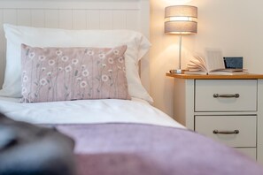 A neatly made bed with a floral pillow, a bedside lamp, and a nightstand with a book and a small electronic device. - 3 Bed in Callington (oc-t46349) (Harrowbarrow)