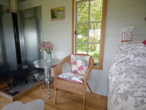 A cozy room with a fireplace, a wicker chair with a floral cushion, a bed with floral bedding, and a window with a view of greenery. - 1 Bed in Abergavenny (oc-42846) (Llangenny)