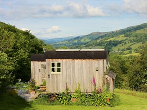 A rustic wooden structure with a window and a door, surrounded by lush greenery and hills in the background. - 1 Bed in Abergavenny (oc-42846) (Llangenny)