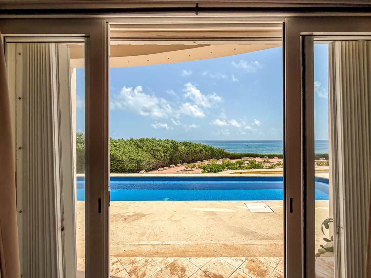 Blick durch eine Schiebetür auf einen Poolbereich mit blauem Himmel und dem Meer im Hintergrund.