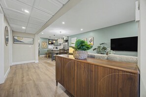 A modern kitchen with white cabinets, a dining area with wooden furniture, and a living room with a flat-screen TV.