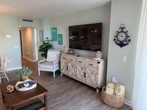A living room with a wooden cabinet, a white armchair, a dining table with a basket, and a wall-mounted clock. - Relaxing Oceanview Beach Retreat in Ocean Creek (Myrtle Beach)