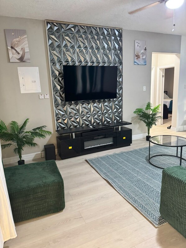Een moderne woonkamer met een flatscreen televisie aan een wand met een geometrisch patroon, een glazen salontafel en twee groene poefjes.