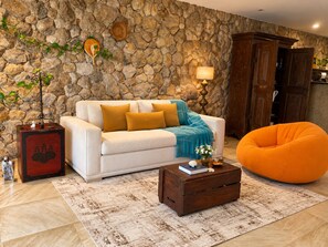 A living room with a stone wall, a white sofa with yellow cushions, a wooden cabinet, and an orange bean bag. - Spacious loft at the foot of Pedra da Gávea.  Gated community with 24-hour security. (Rio de Janeiro)