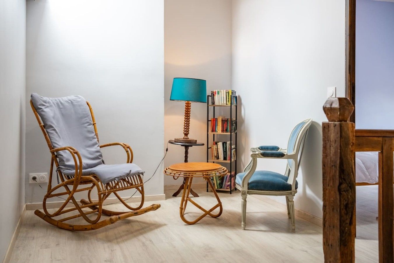 Une pièce comprenant un fauteuil à bascule en osier, une petite table ronde, une bibliothèque et une lampe.
