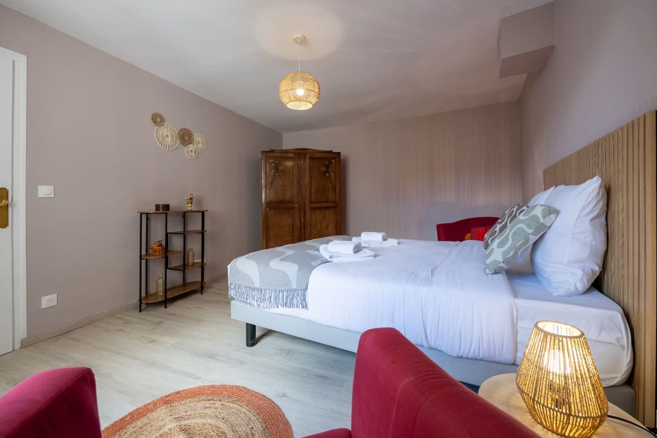 Una habitació d'hotel amb un llit, un armari, una prestatgeria plena d'objectes, una cadira vermella i una làmpada.