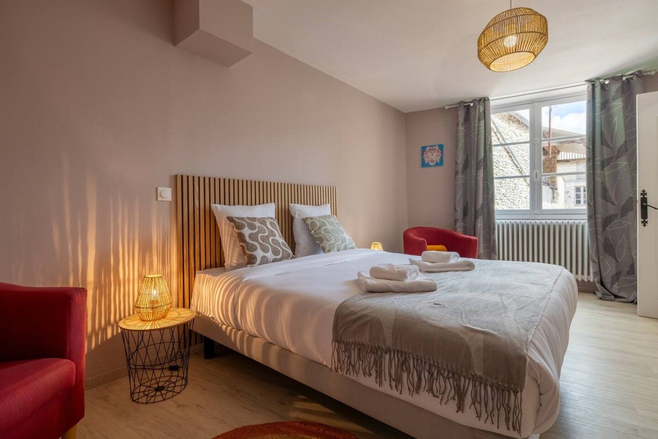 A modern bedroom with a large bed, a red chair, a small table with a lamp, and a view of the outside through a window.
