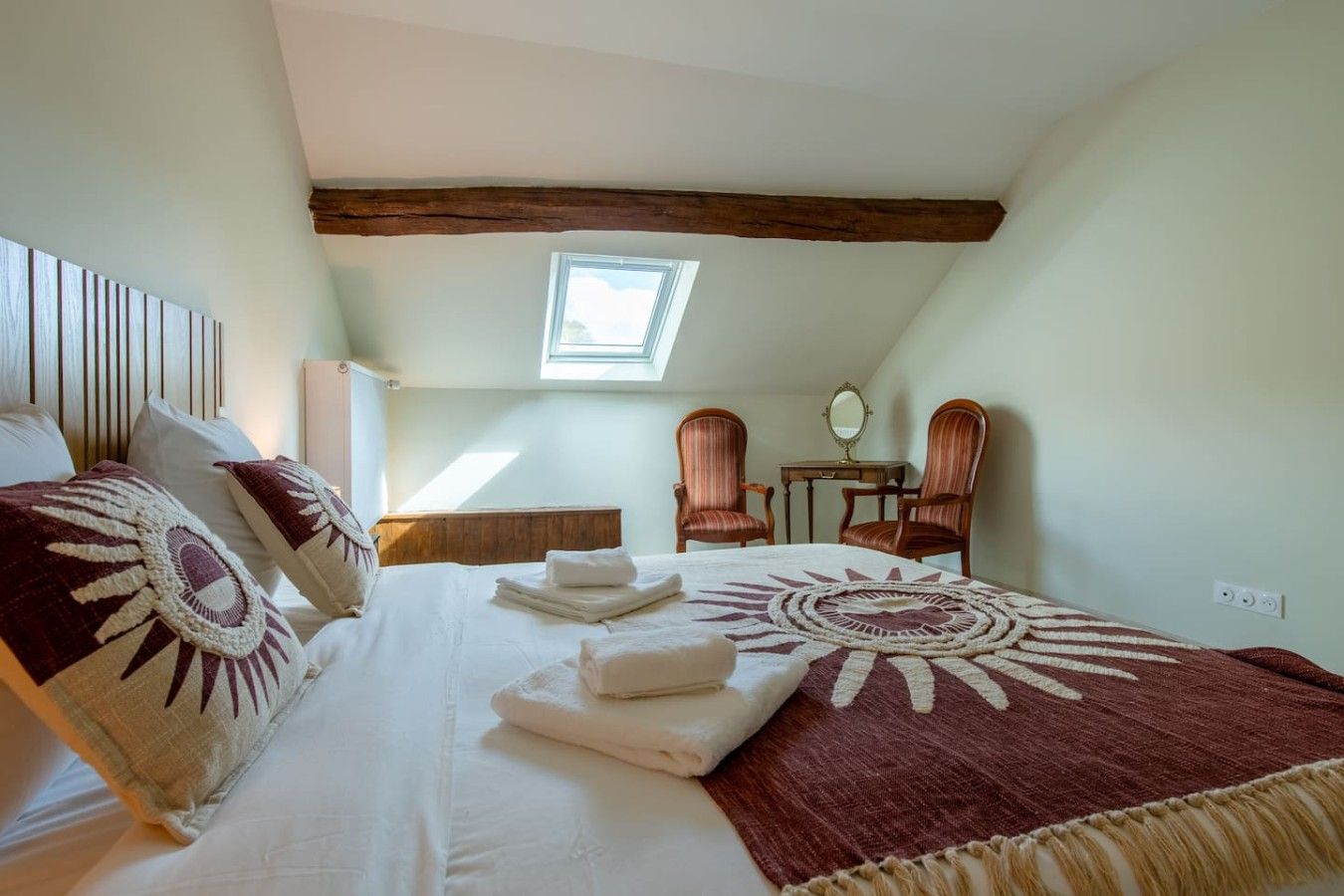 Un dormitorio con cama, dos sillas, una mesita, un espejo y un tragaluz.