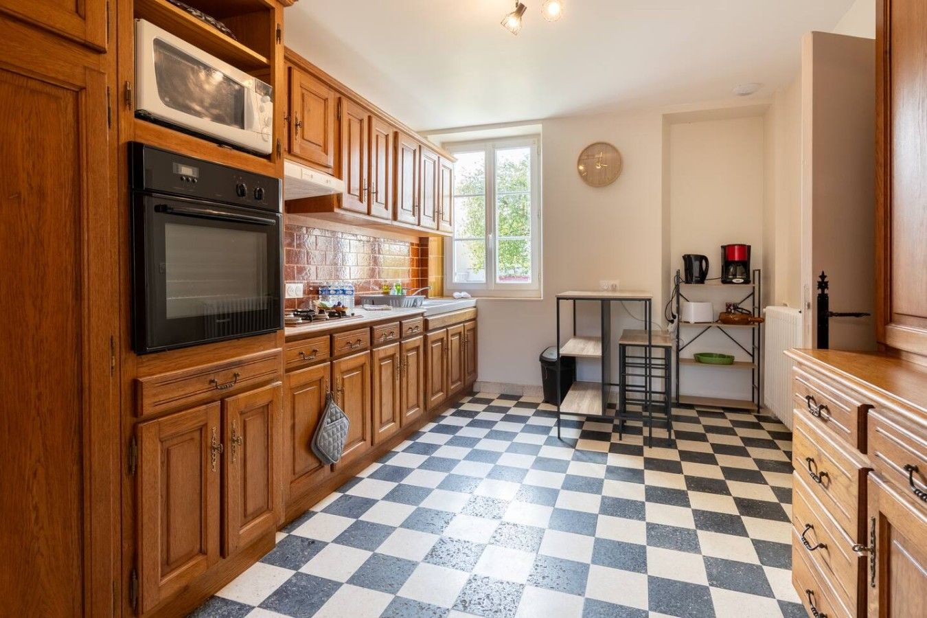 Una cocina con armarios de madera, un horno negro, un microondas, piso a cuadros y una ventana con vista a follaje.