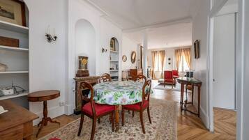 A dining area with a round table, chairs, and a floral tablecloth. There are wooden chairs with red cushions, a wooden side table, and a small round table with a lamp. The room has white walls, wooden floors, and a red rug.