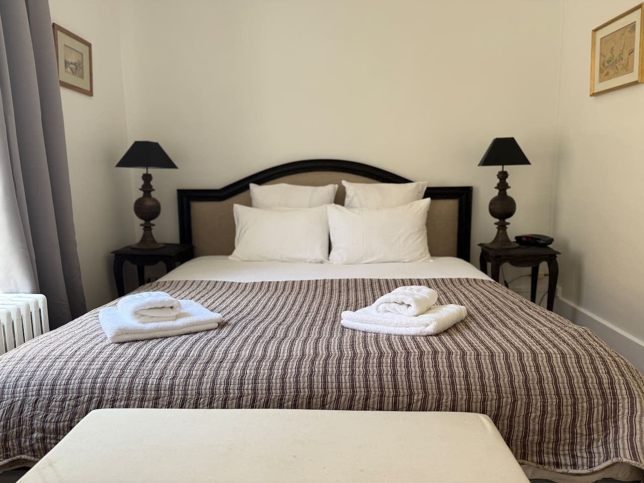 Una habitación de hotel con cama, dos lámparas de noche, un radiador y una ventana con cortinas.