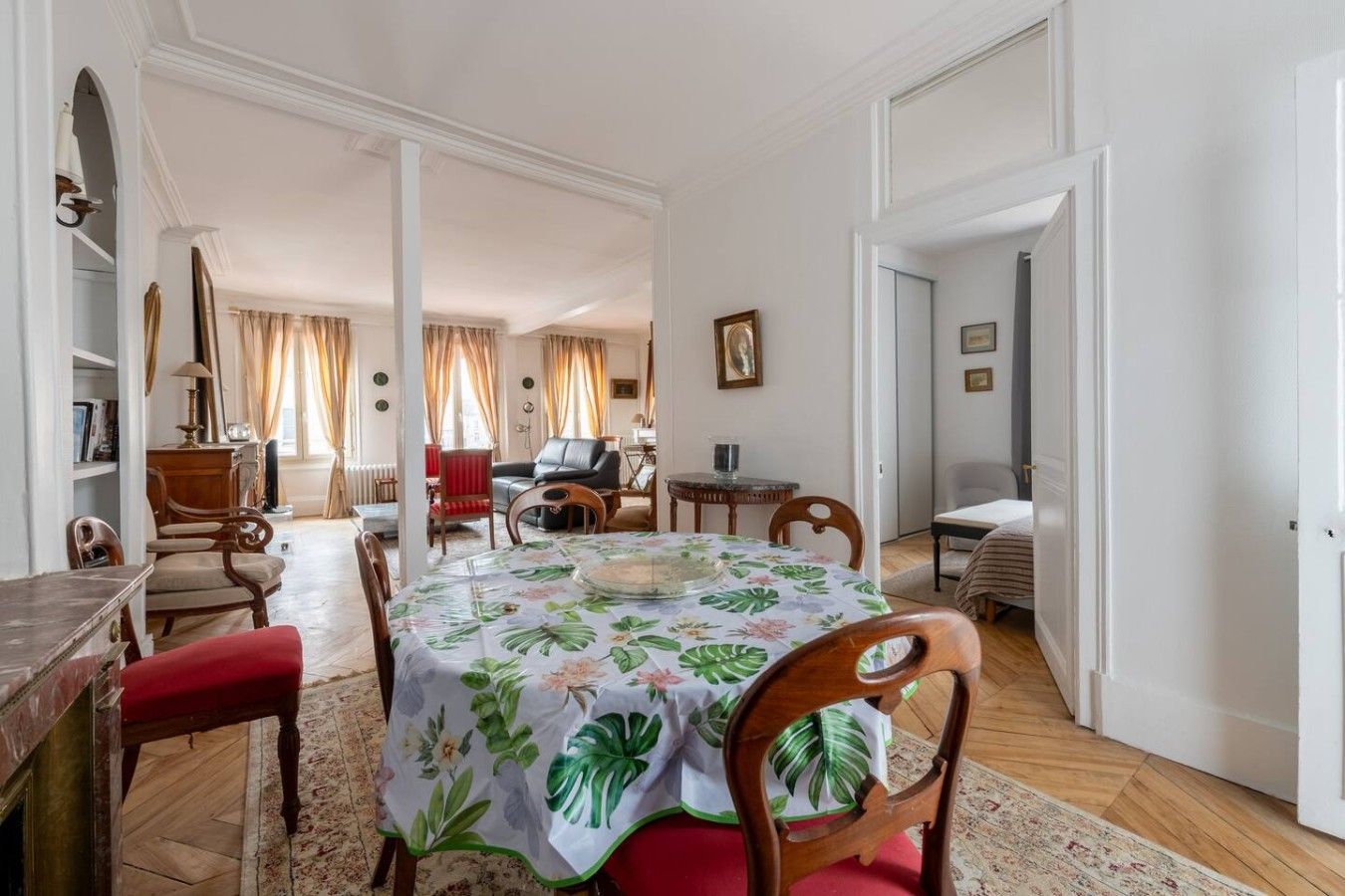 A dining area with a round table covered by a floral tablecloth, surrounded by wooden chairs.