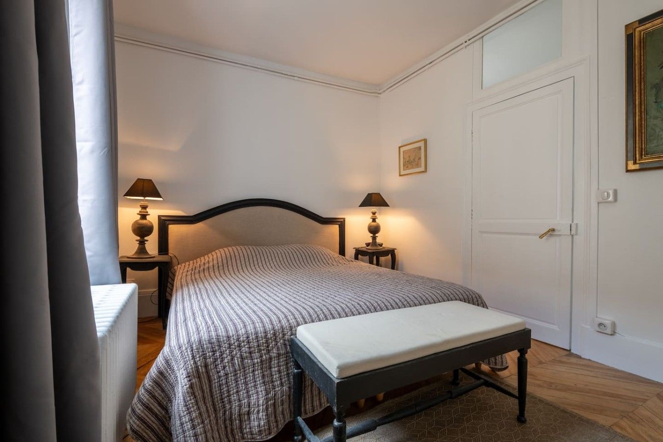 Een slaapkamer met een bed, nachtkastjes, een bankje, een deur en een ingelijst kunstwerk aan de muur.