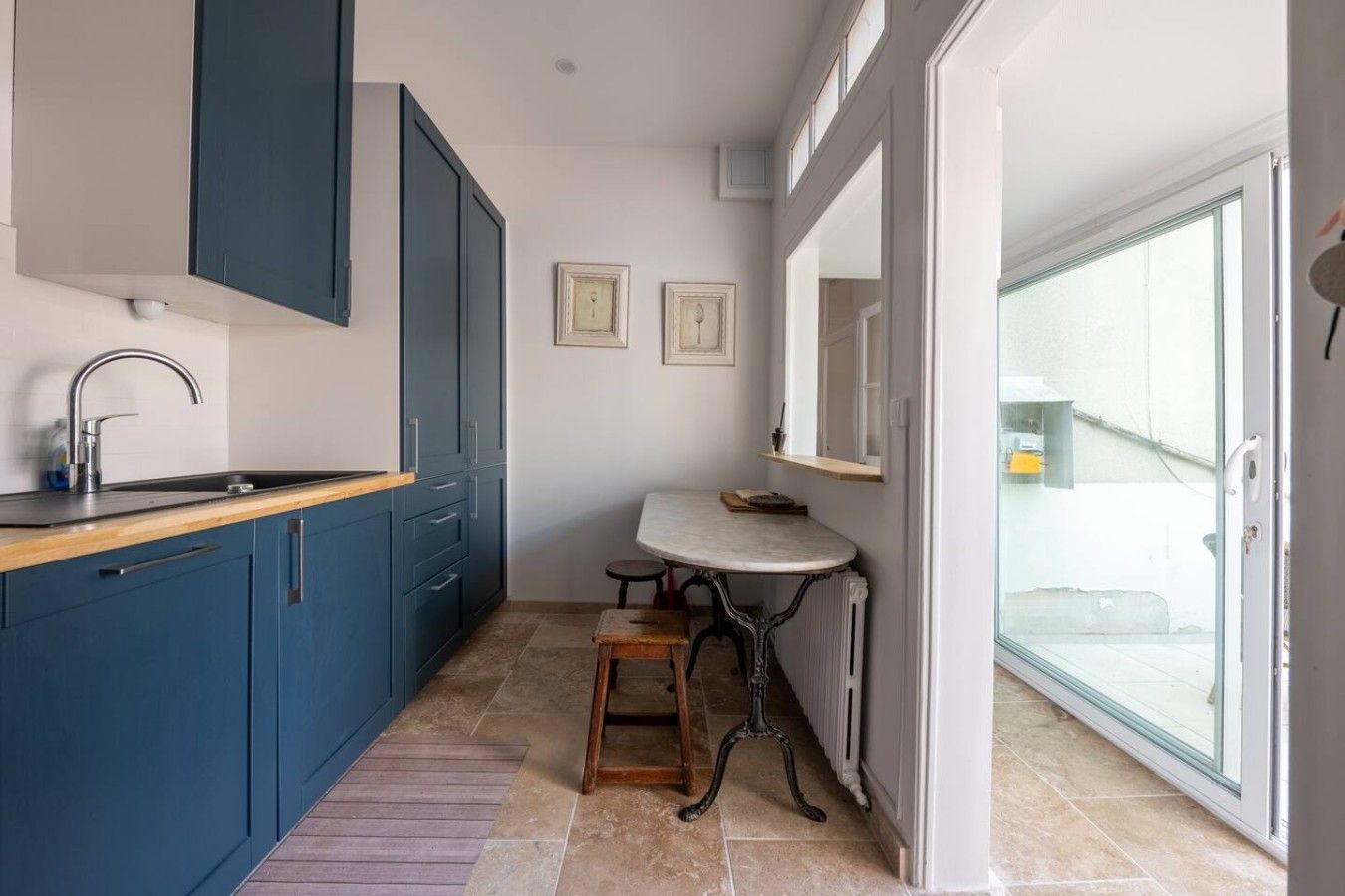 A narrow kitchen with blue cabinets, a sink, and a small table with a stool.