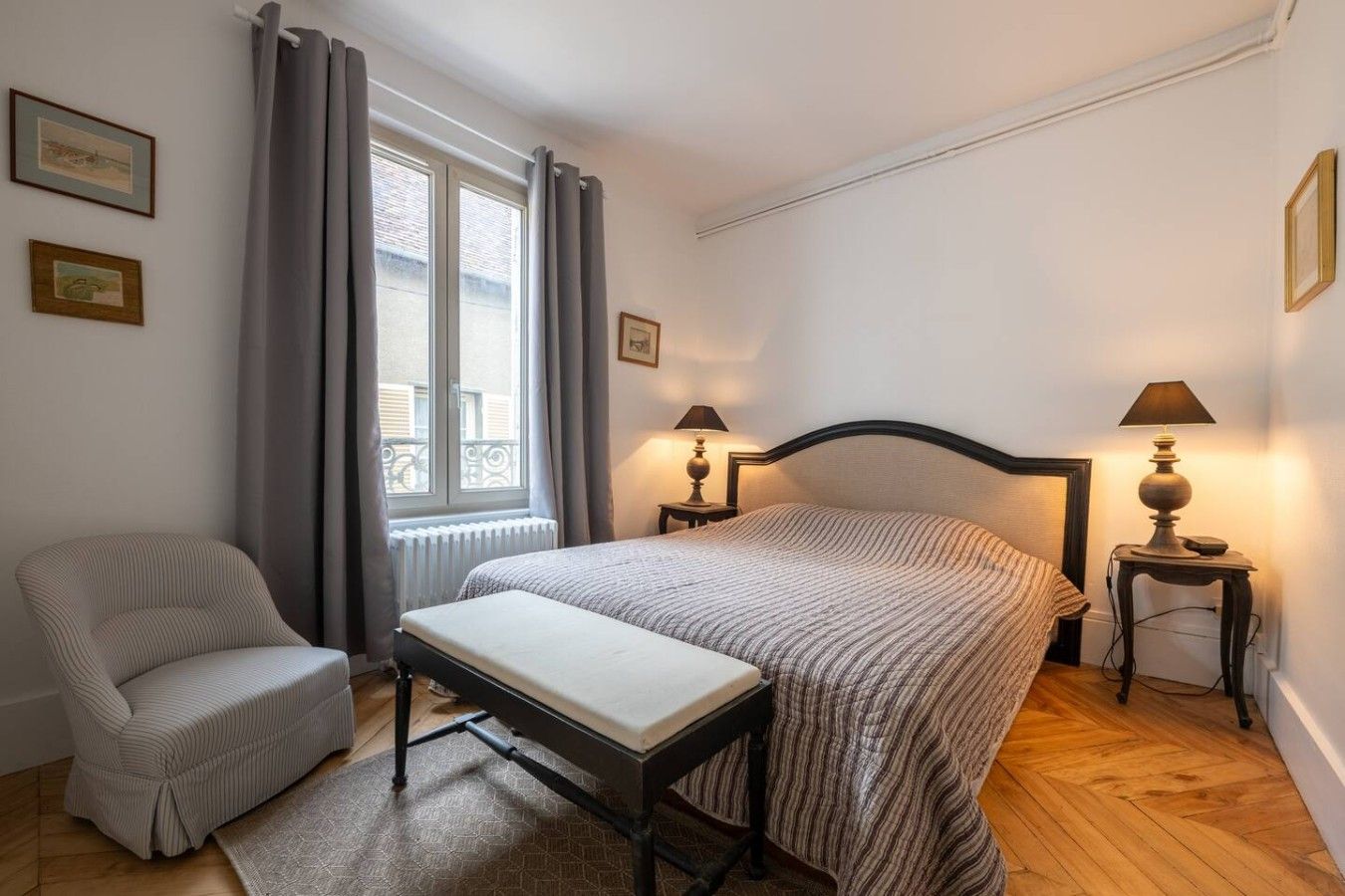 Een slaapkamer met een bed, een stoel, twee nachtkastjes met lampen, een raam met gordijnen en een radiator.