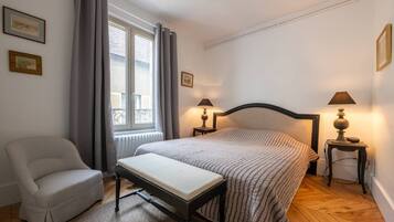 Een slaapkamer met een bed, een stoel, twee nachtkastjes met lampen, een raam met gordijnen en een radiator.