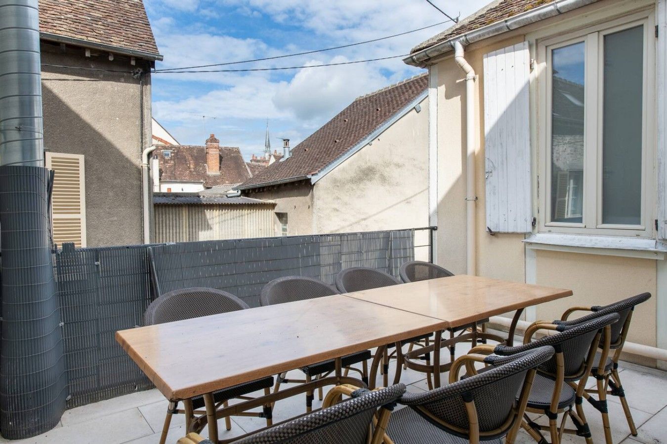 An outdoor patio with a wooden table and wicker chairs, a metal fence, and a view of neighboring buildings.