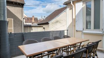 An outdoor patio with a wooden table and wicker chairs, a metal fence, and a view of neighboring buildings.