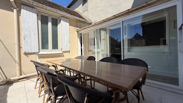 Un espace extérieur aménagé pour les repas, avec une table en bois et des chaises en osier, une porte-fenêtre coulissante et un mur extérieur blanc.
