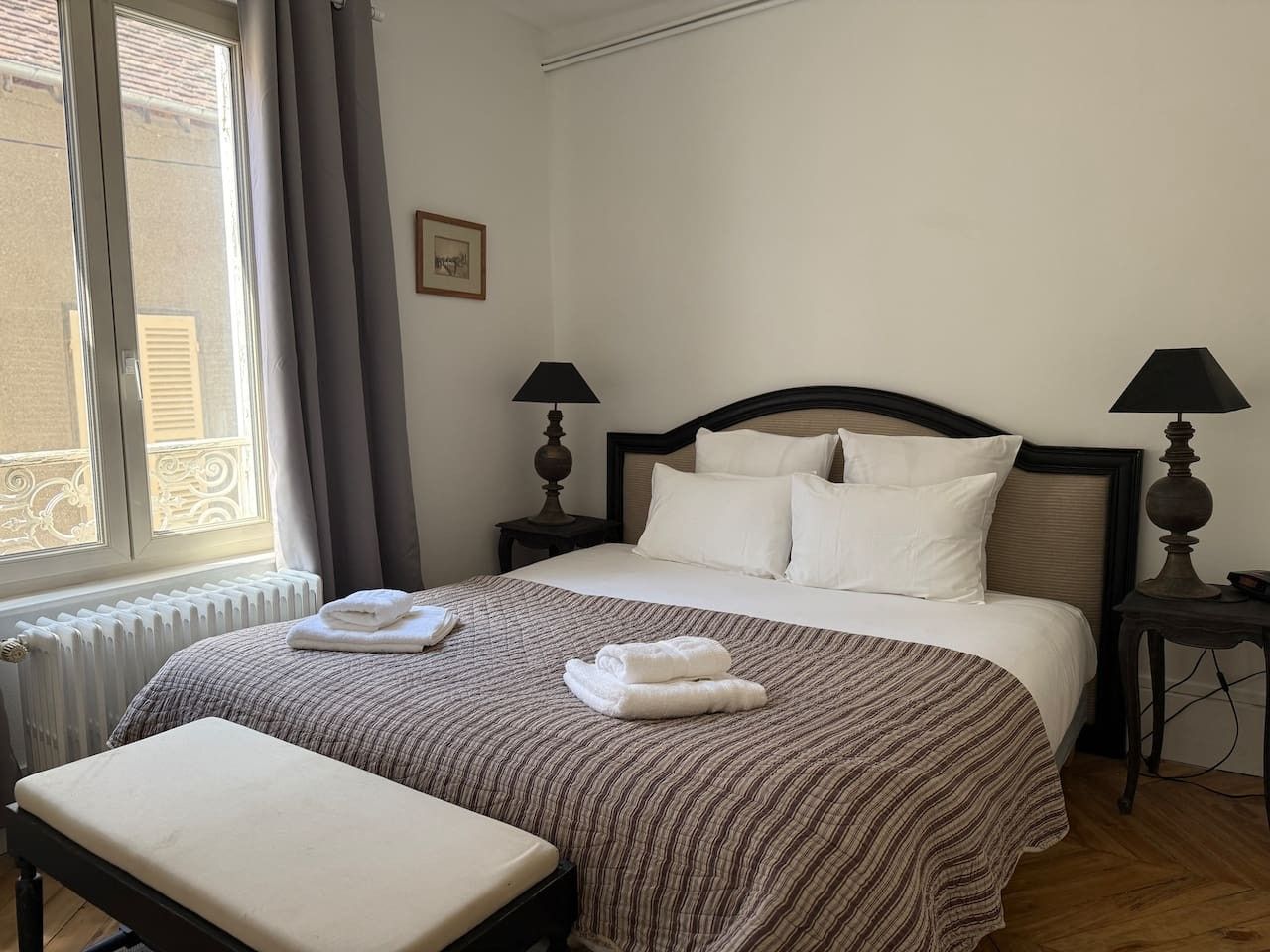 A hotel room with a bed, two bedside tables, a window with curtains, and a radiator.