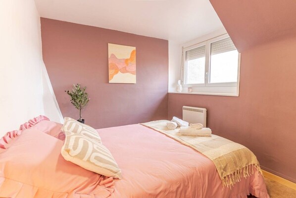 Un dormitorio con una cama, almohadas, una mesita de noche con una planta, un radiador y una ventana.