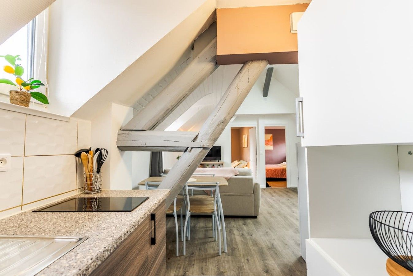 Una cocina moderna con una escalera de madera, una zona de comedor y un dormitorio visible a través de una puerta abierta.