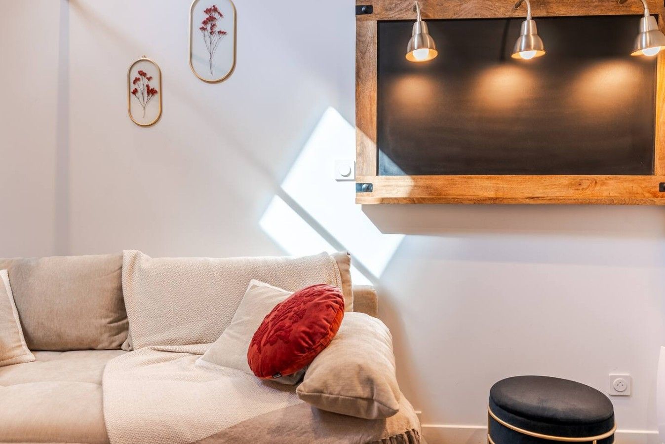 Un salon avec un canapé, un coussin rouge, un petit tabouret et un luminaire fixé au mur.