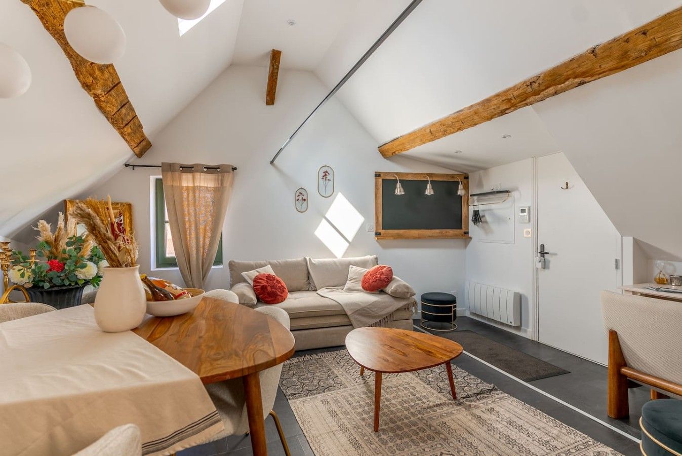 A cozy attic bedroom with a sloped ceiling, a wooden ceiling beam, a bed, a small table, a sofa, and a window with curtains.