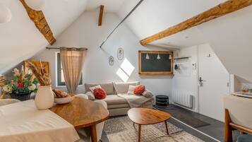 A cozy attic bedroom with a sloped ceiling, a wooden ceiling beam, a bed, a small table, a sofa, and a window with curtains.