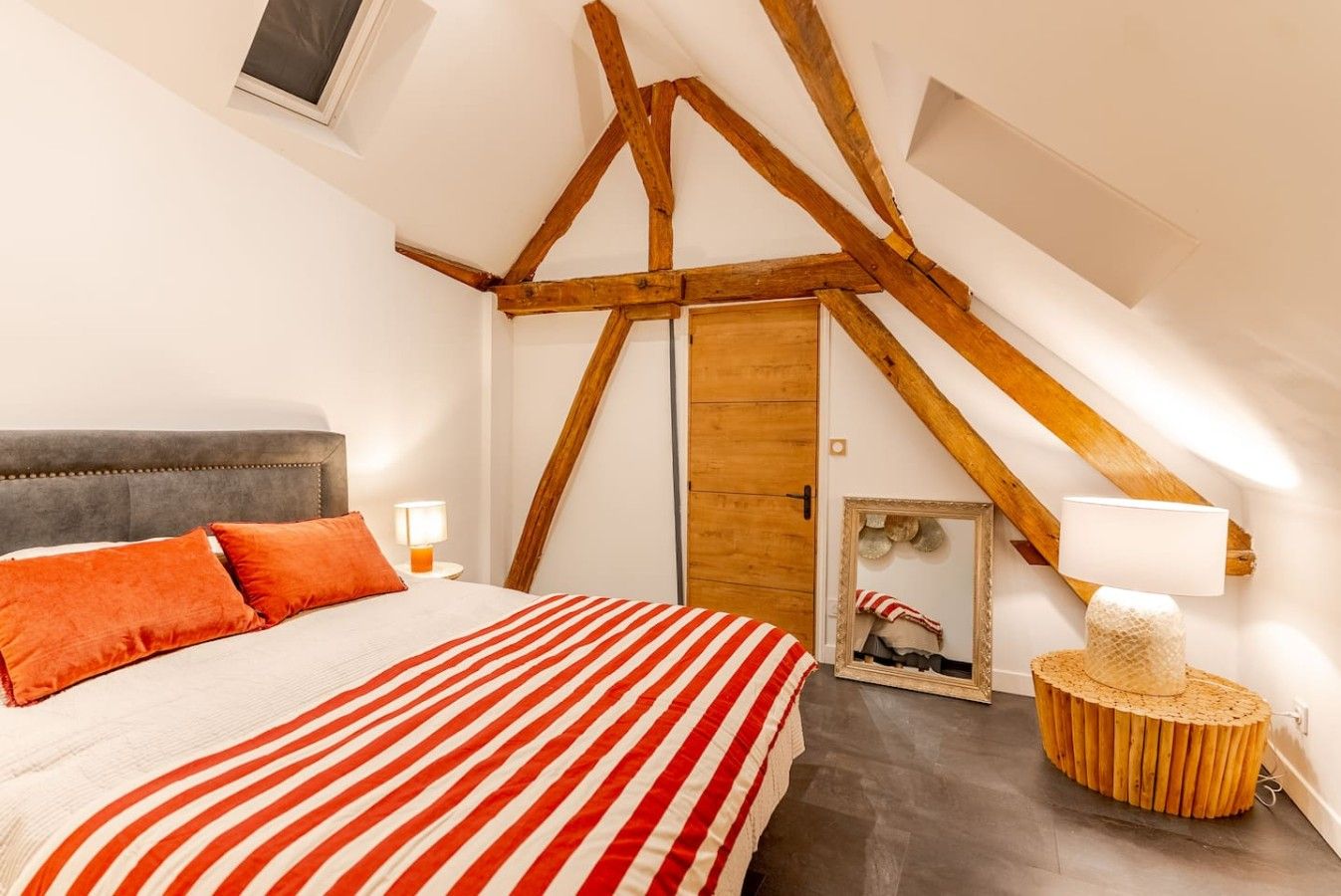 A bedroom with a sloped ceiling, a bed with striped bedding, a wooden headboard, and a round wooden side table with a lamp.