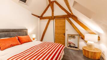 A bedroom with a sloped ceiling, a bed with striped bedding, a wooden headboard, and a round wooden side table with a lamp.