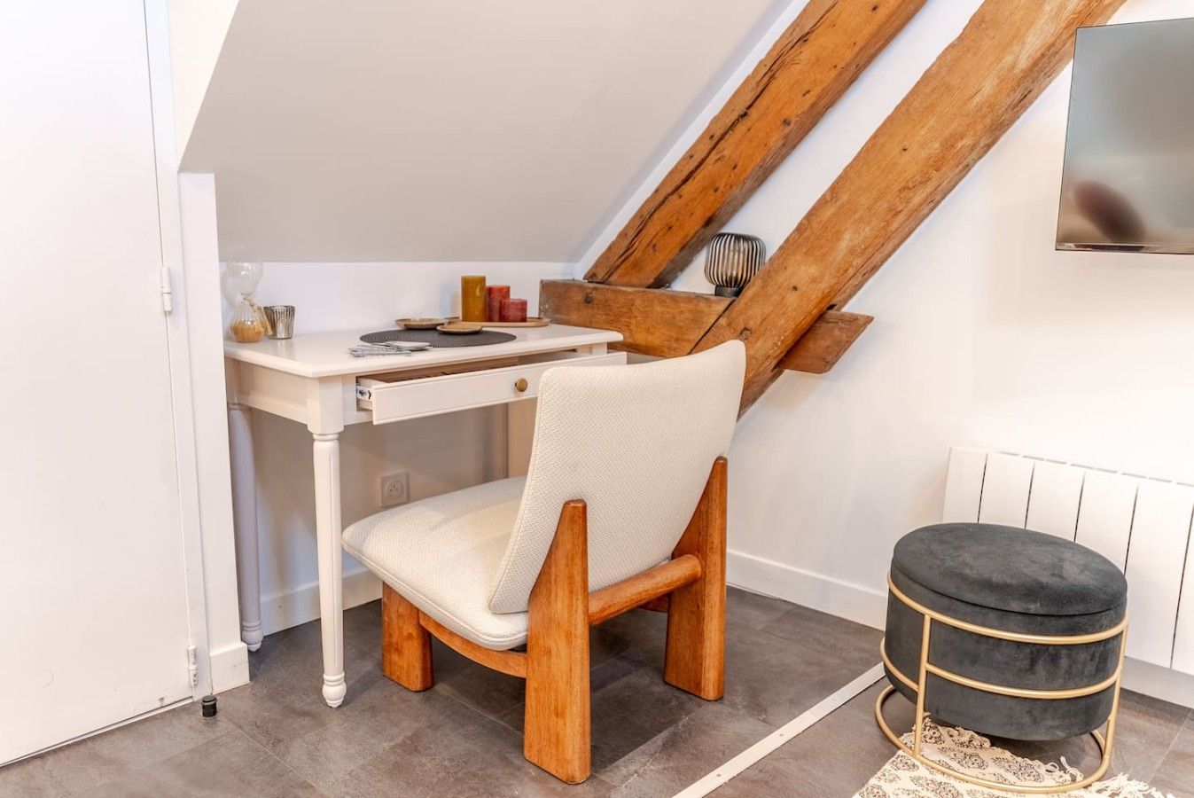 A room with a white desk, a wooden chair, a black ottoman, and a slanted ceiling.