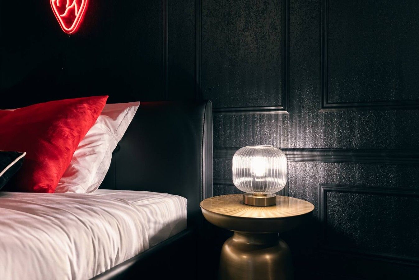 A bedroom with a bed, a round nightstand with a lamp, and a neon heart sign on the wall.