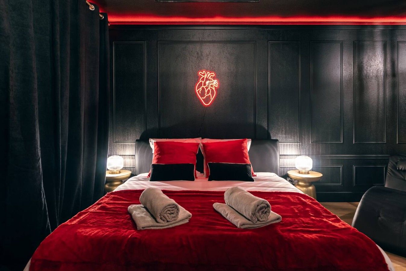 Une chambre d’hôtel avec un grand lit, une literie rouge, un cœur lumineux accroché au mur et deux lampes de chevet.