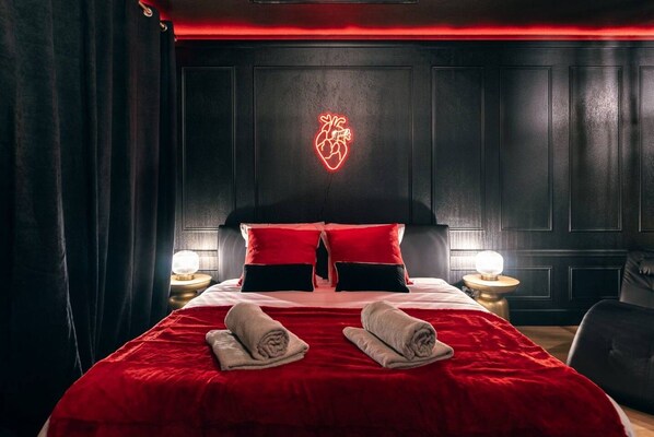 A hotel room with a large bed, red bedding, a neon heart sign on the wall, and two bedside lamps.