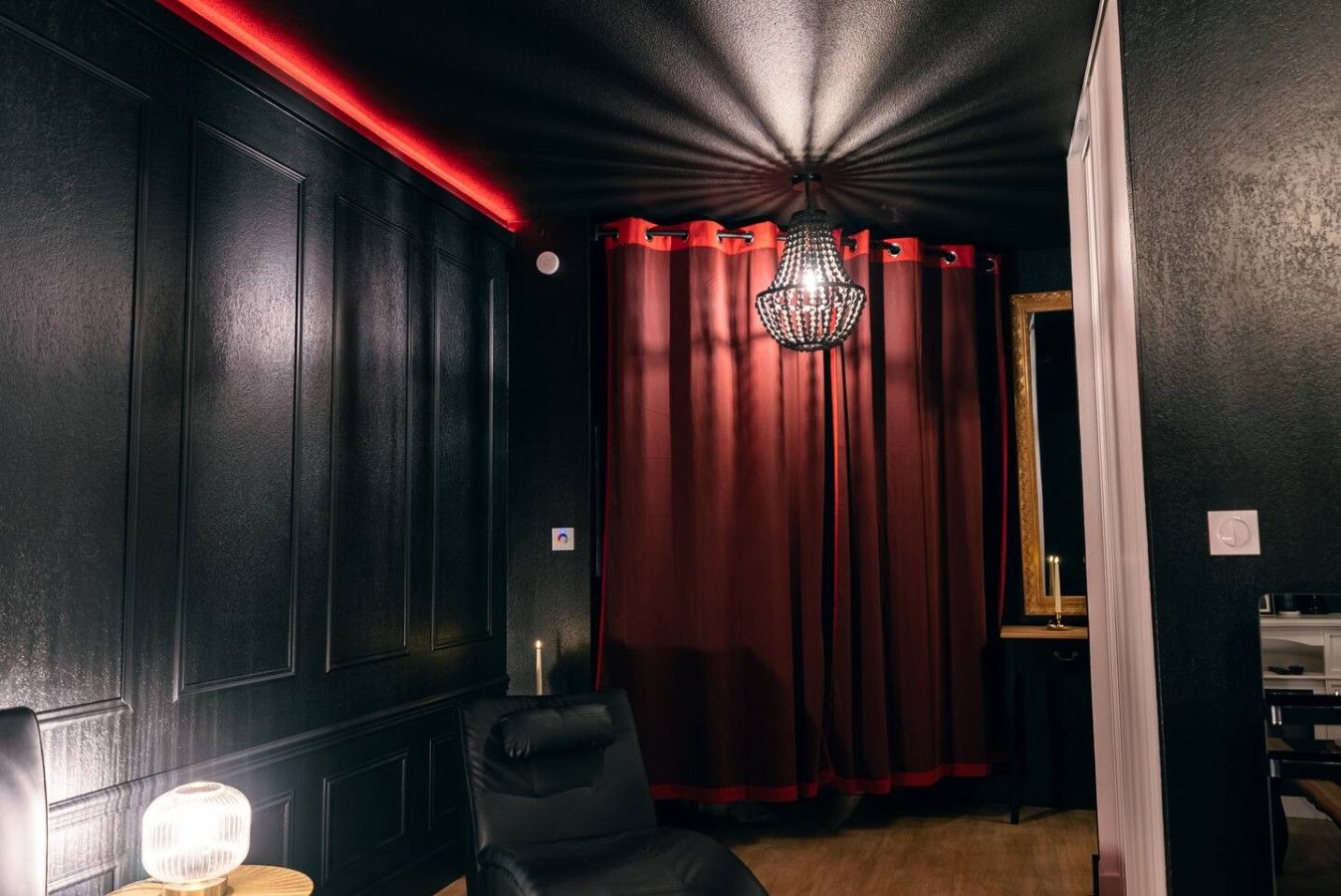 A room with dark walls, a red curtain, a chandelier, and a black leather chair.