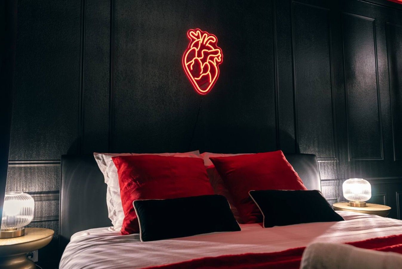 Une chambre à coucher avec un lit, des oreillers rouges et noirs, un néon en forme de cœur accroché au mur et des lampes de chevet.