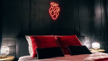 Une chambre à coucher avec un lit, des oreillers rouges et noirs, un néon en forme de cœur accroché au mur et des lampes de chevet.