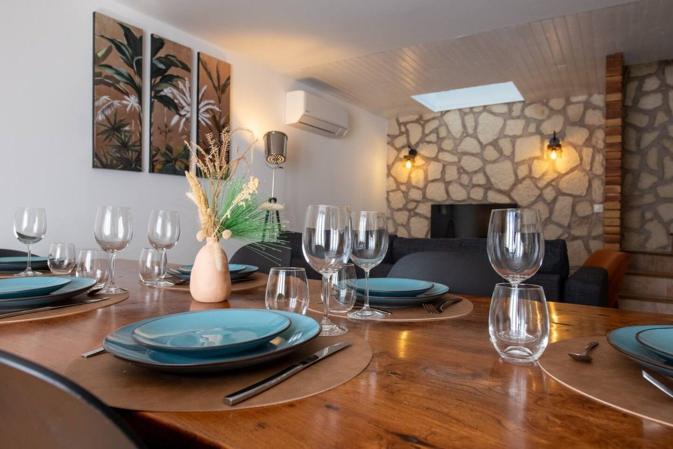 Un coin repas avec une table en bois dressée pour un repas, entourée de chaises. On y trouve deux luminaires fixés au mur et un mur en pierre qui sert d’élément décoratif.