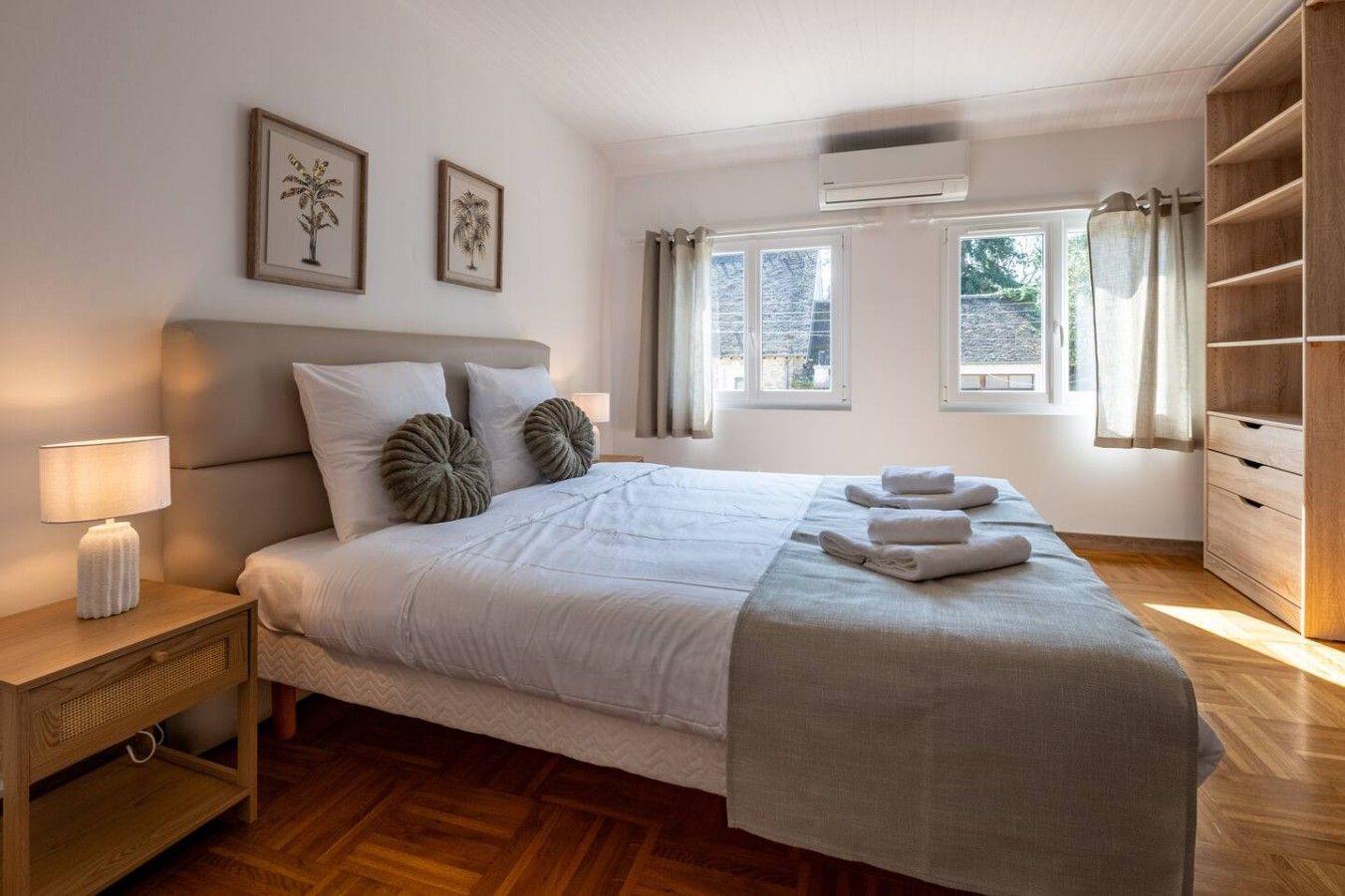 A bedroom with a bed, bedside table, lamp, air conditioning unit, windows, and a wardrobe.