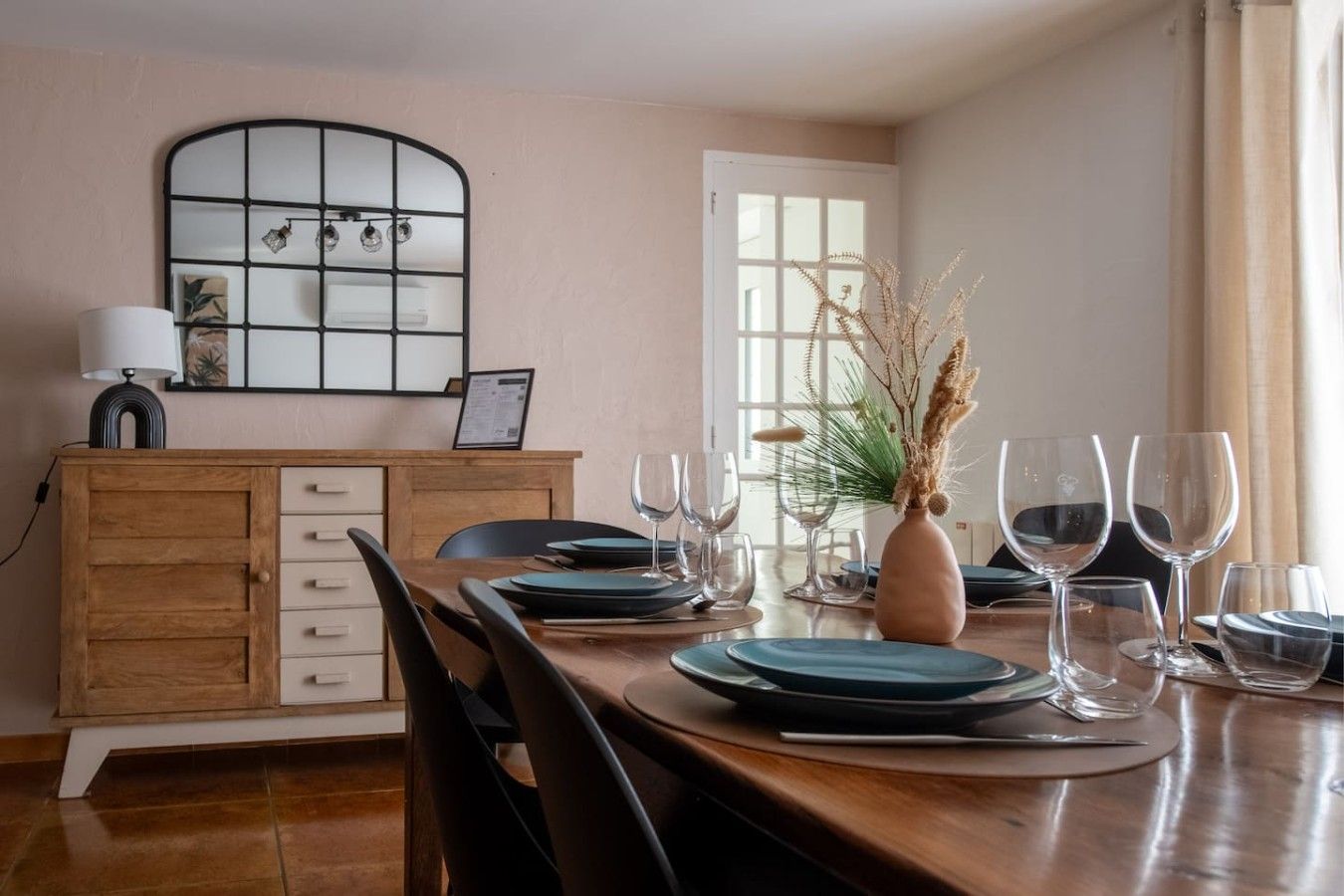 Ein Essbereich mit einem Holztisch, gedeckt für vier Personen, einer Vase mit Trockenpflanzen und einem Spiegel über dem Tisch.