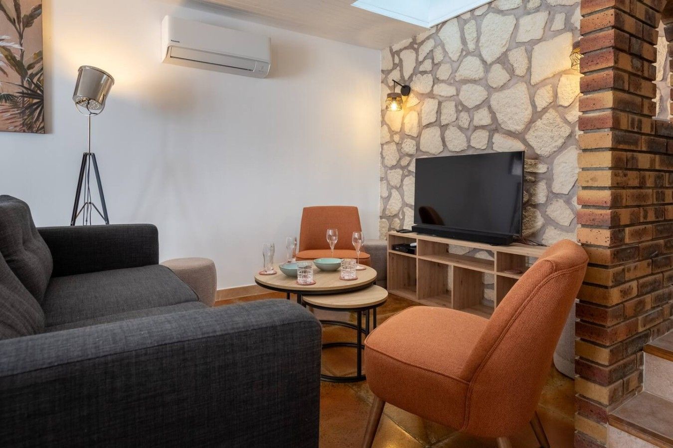 A living room with a sofa, a chair, a small round table with glasses, a flat-screen TV, and a stone wall.