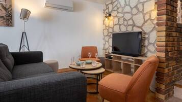 A living room with a sofa, a chair, a small round table with glasses, a flat-screen TV, and a stone wall.