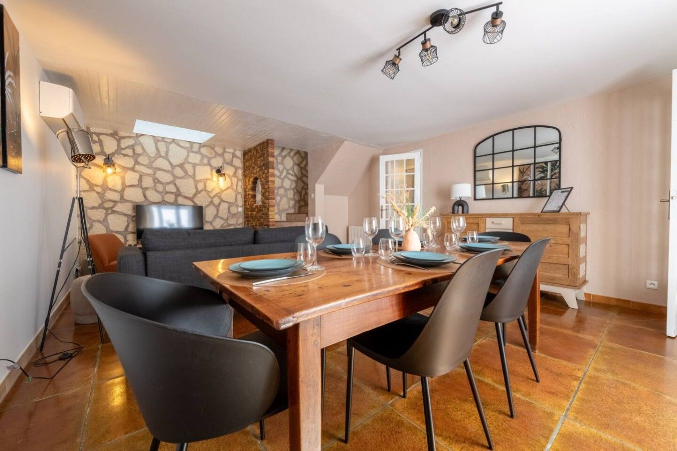 A dining area with a wooden table set for six, surrounded by black chairs, a stone wall, and a modern light fixture above.