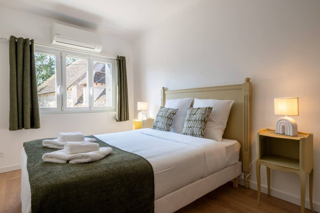 Een netjes opgemaakt bed met een groene deken en witte kussens, een nachtkastje met een lamp, en een raam met gordijnen.