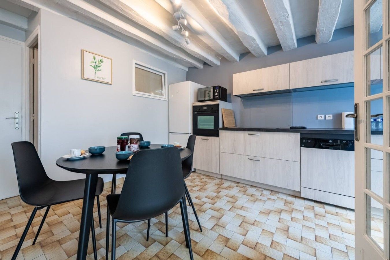 A modern kitchen with a dining area, featuring a herringbone floor pattern, light-colored cabinetry, and black chairs.