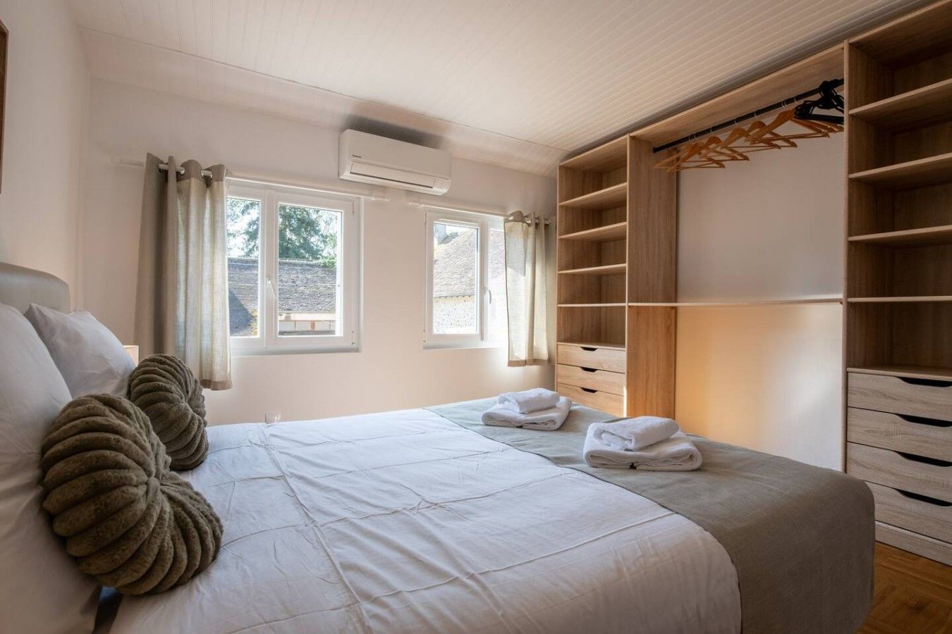 A bedroom with a large bed, two windows, an air conditioning unit, and a walk-in closet with wooden shelves and drawers.