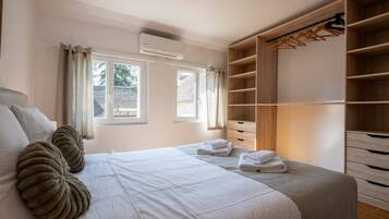 A bedroom with a large bed, two windows, an air conditioning unit, and a walk-in closet with wooden shelves and drawers.
