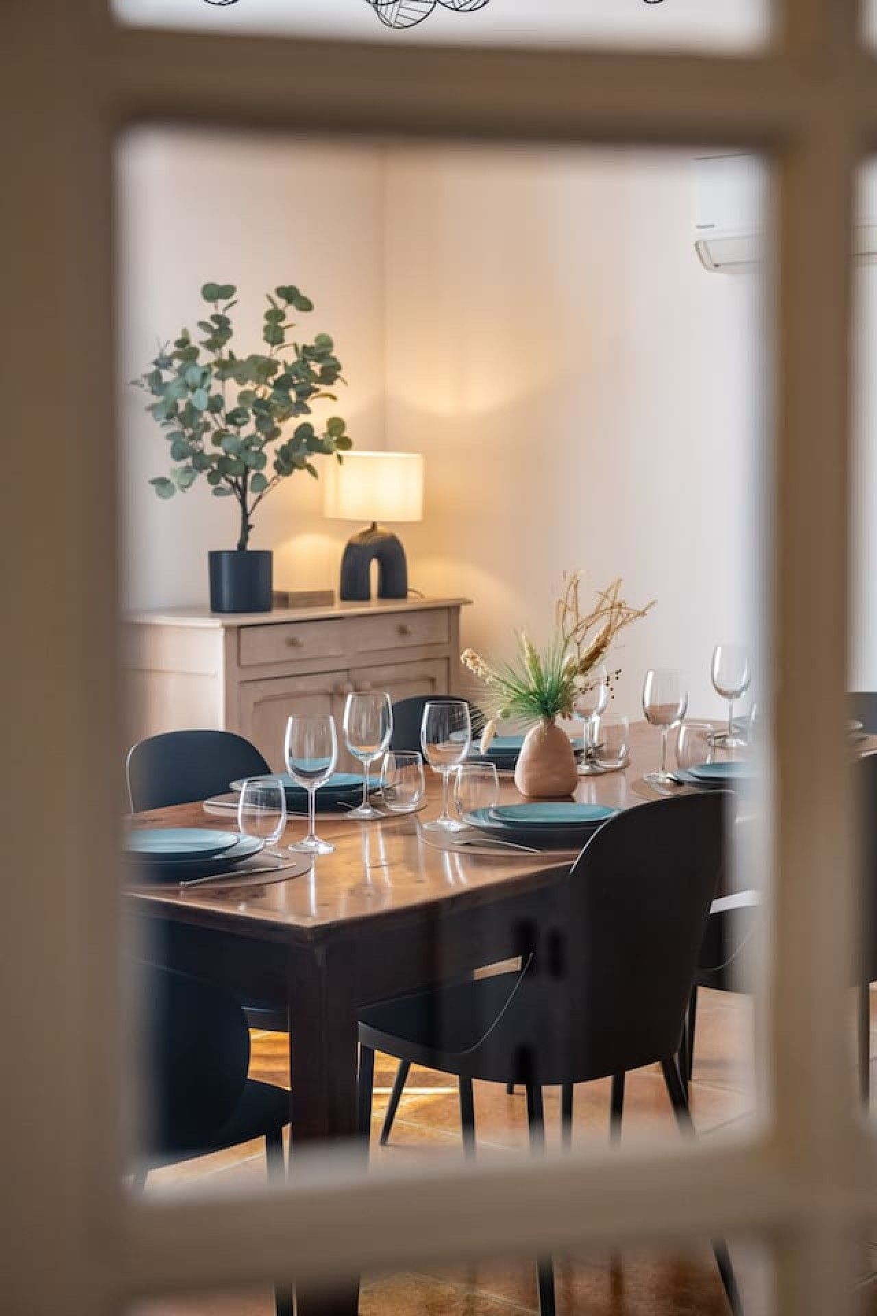 Un coin repas avec une table dressée pour quatre personnes, des chaises, une plante en pot et une lampe.